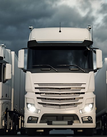 Camion Blanc Moderne Face à un Ciel d'Orage Gros plan frontal sur un camion blanc moderne avec phares allumés, flanqué d'autres remorques, sous un ciel sombre et menaçant.